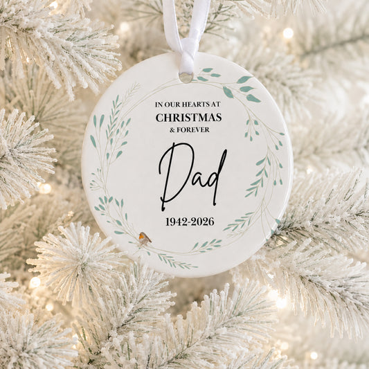 A round white Christmas ornament hanging on a frosted white Christmas tree branch, with printed black text reading 'IN OUR HEARTS AT CHRISTMAS & FOREVER Dad 1942-2026' and a small green botanical wreath design.