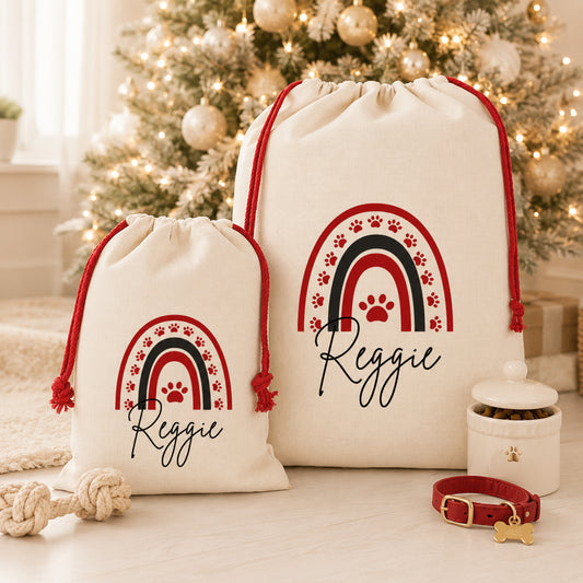 Two cream drawstring gift bags with red and black rainbow paw-print designs and the name 'Reggie' printed on them, styled on a tabletop in front of a decorated Christmas tree with a pet collar and treat jar nearby.