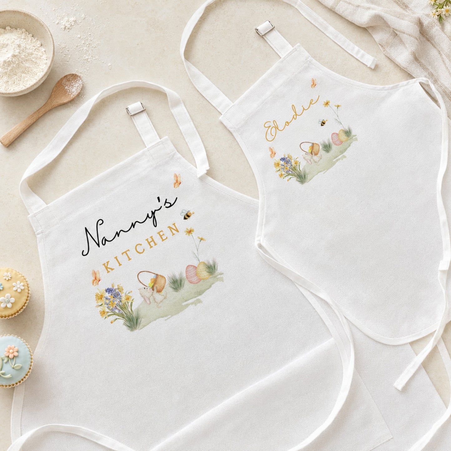 Two white fabric aprons laid flat on a light beige surface, each with embroidered Easter-themed artwork and personalized text reading 'Nanny's Kitchen' and 'Eddie'. The aprons are styled with baking props including flour, a wooden spoon, cupcakes, and a cloth.