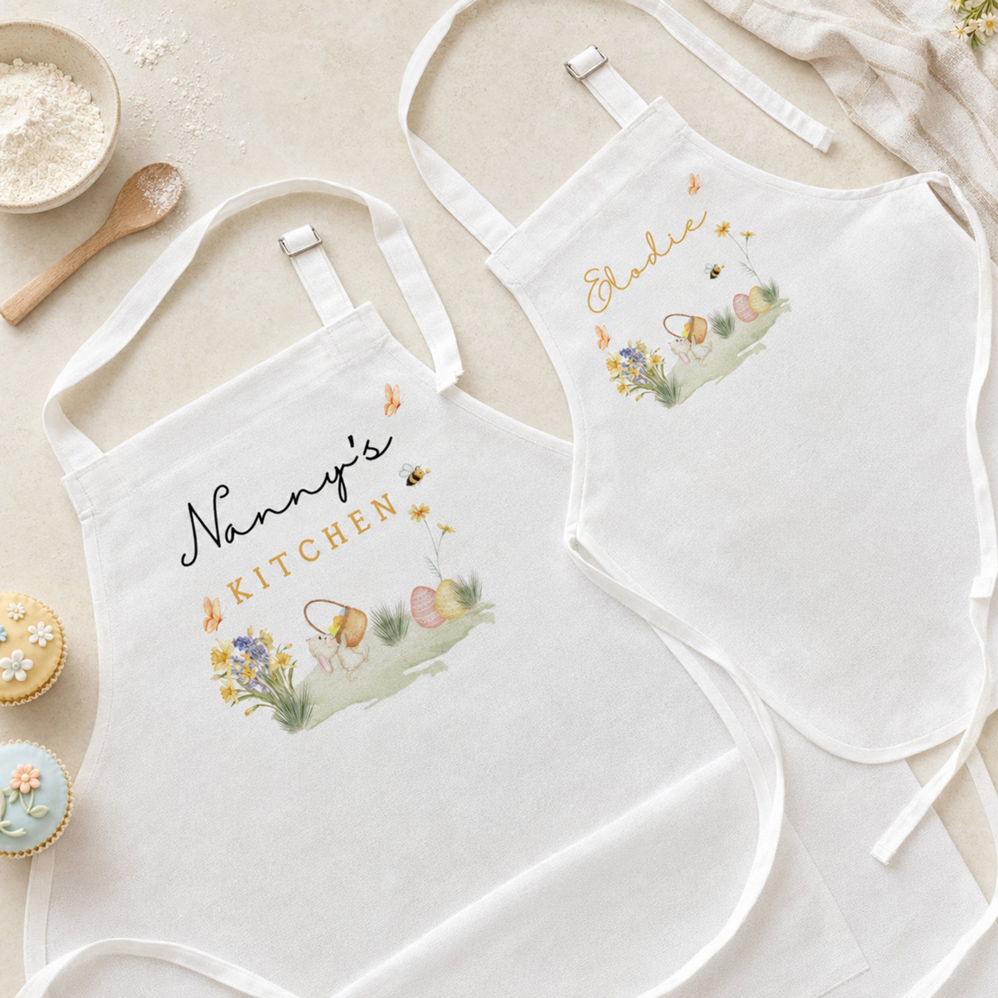 Two white fabric aprons laid flat on a light beige surface, each with embroidered Easter-themed artwork and personalized text reading 'Nanny's Kitchen' and 'Eddie'. The aprons are styled with baking props including flour, a wooden spoon, cupcakes, and a cloth.