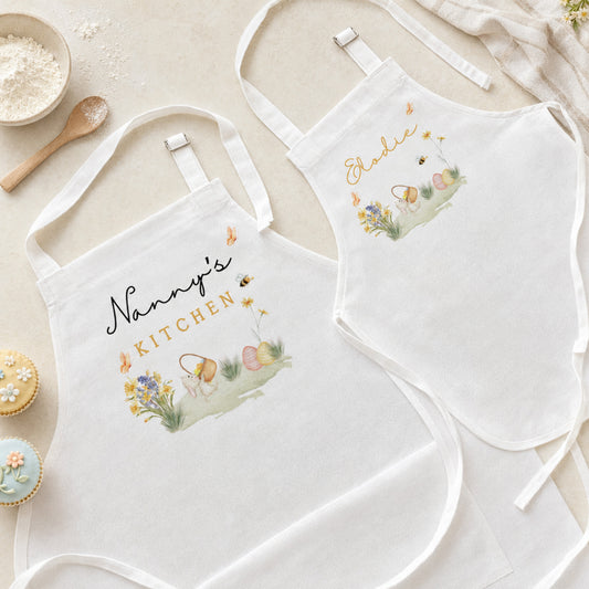 Two white fabric aprons laid flat on a light beige surface, each with embroidered Easter-themed artwork and personalized text reading 'Nanny's Kitchen' and 'Eddie'. The aprons are styled with baking props including flour, a wooden spoon, cupcakes, and a cloth.