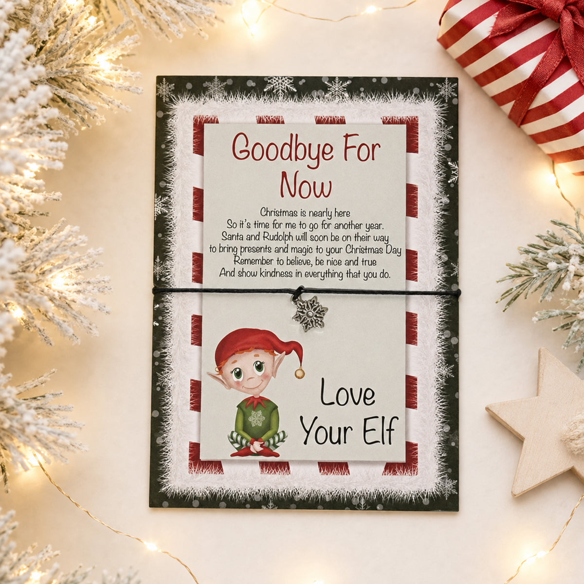 A Christmas-themed farewell card for an elf,  and displayed on a light beige surface with festive props including tinsel, fairy lights, a wrapped gift, and a wooden star.