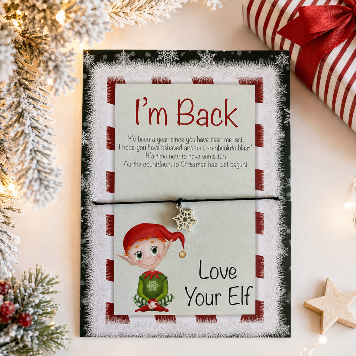 A festive Christmas greeting card with a pull-tab design that reads 'I'm Back' and 'Love Your Elf,' styled on a tabletop with holiday decorations, a wrapped gift, and string lights.