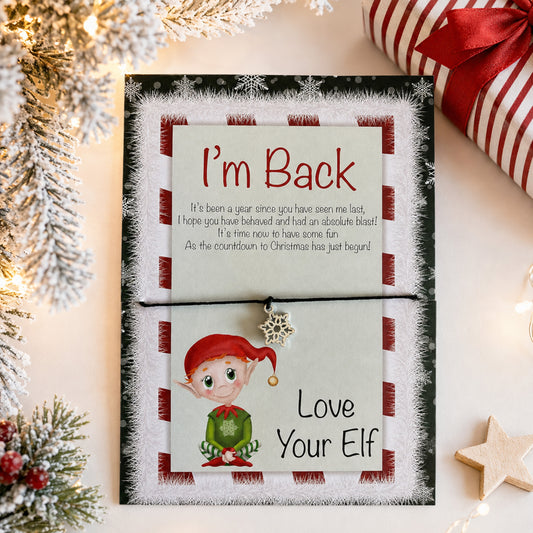 A festive Christmas greeting card with a pull-tab design that reads 'I'm Back' and 'Love Your Elf,' styled on a tabletop with holiday decorations, a wrapped gift, and string lights.