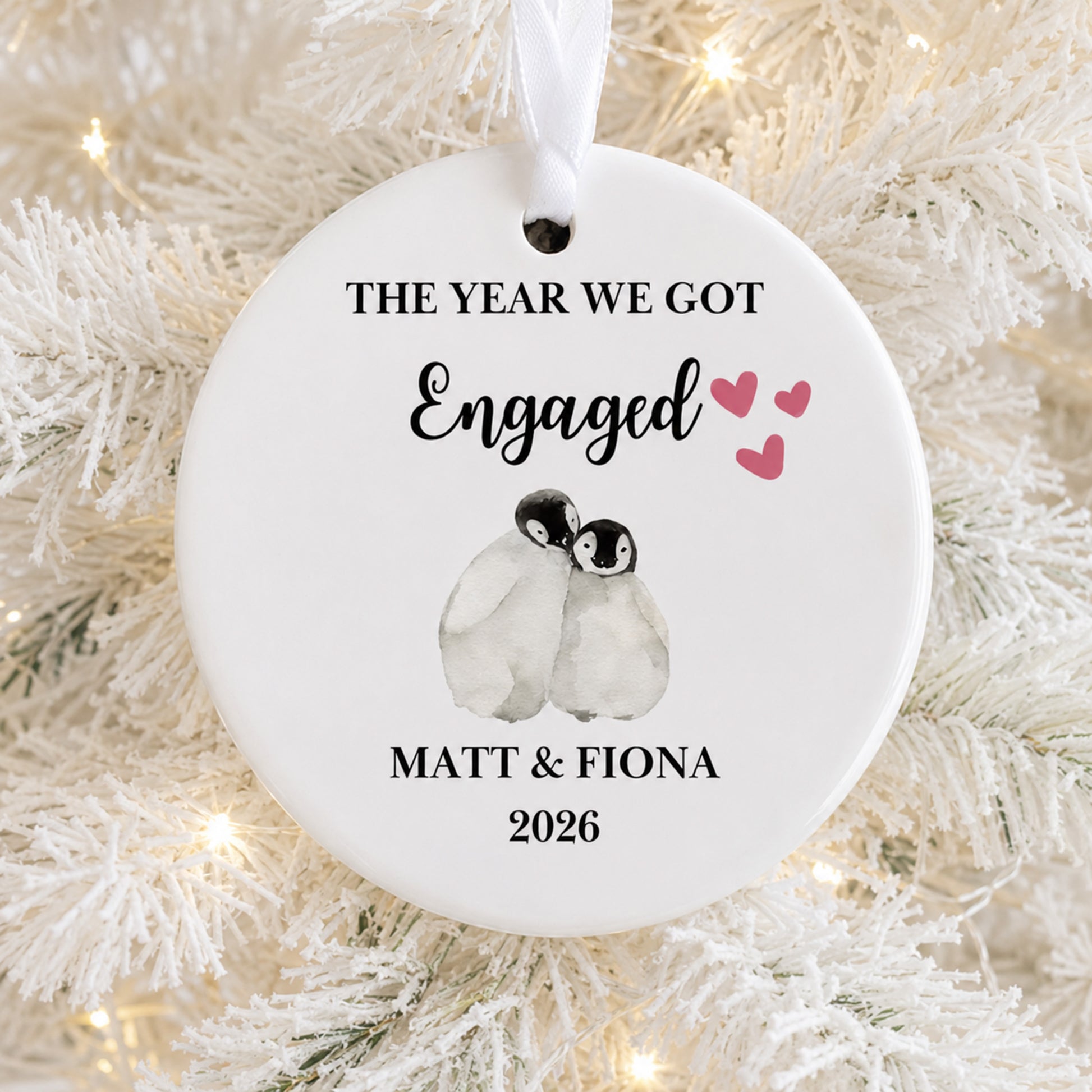 A round white ceramic Christmas ornament hanging on a white frosted Christmas tree branch with warm string lights. The ornament is personalized with the text 'THE YEAR WE GOT Engaged Matt & Fiona 2026' and features a watercolor illustration of two penguins with three pink hearts.