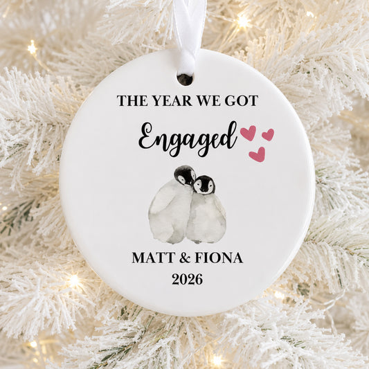 A round white ceramic Christmas ornament hanging on a white frosted Christmas tree branch with warm string lights. The ornament is personalized with the text 'THE YEAR WE GOT Engaged Matt & Fiona 2026' and features a watercolor illustration of two penguins with three pink hearts.