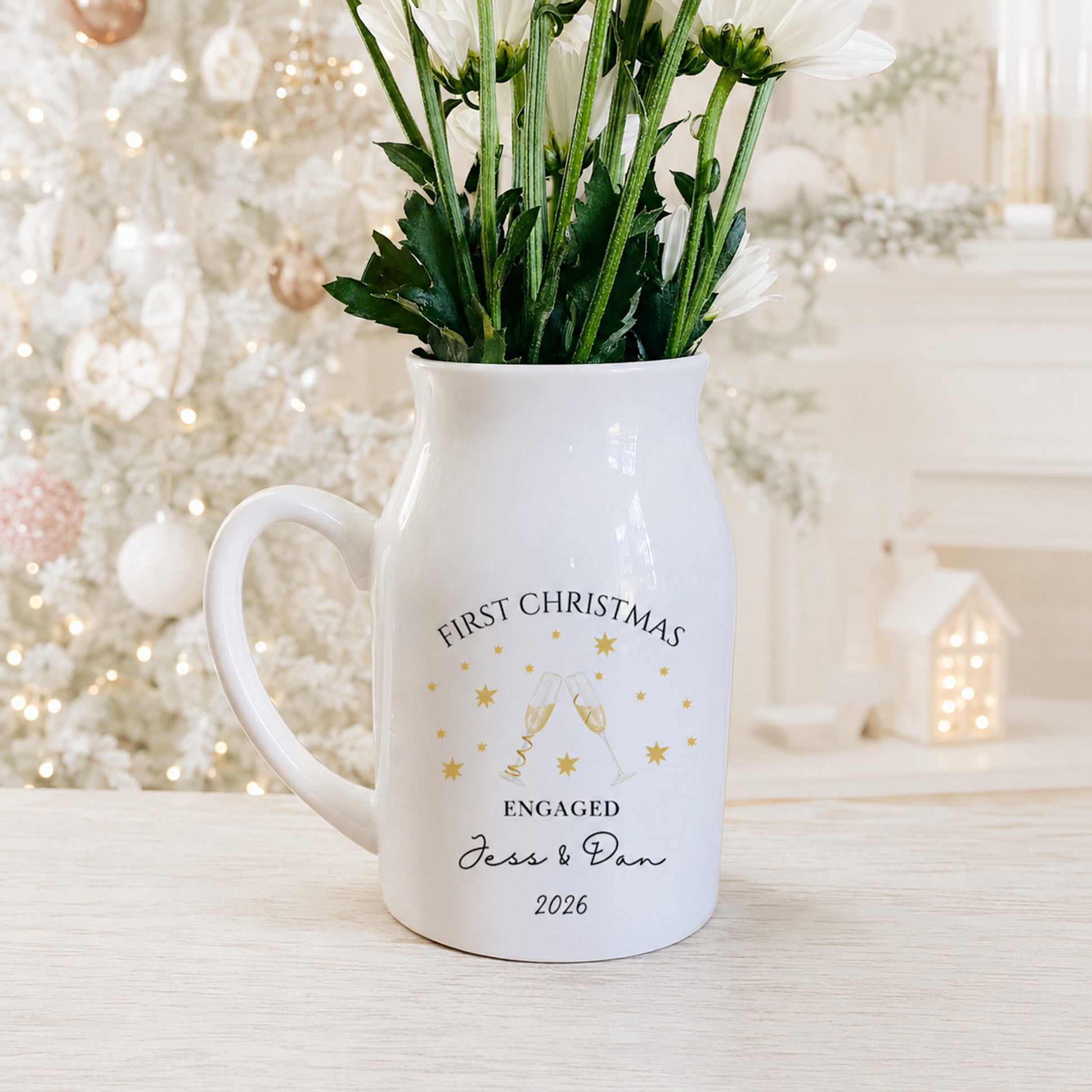 A white ceramic vase shaped like a milk jug, filled with white flowers, sitting on a light wooden table. The mug is personalized with gold foil text reading 'FIRST CHRISTMAS ENGAGED Jess & Dan 2026' and features an illustration of two clinking champagne glasses surrounded by stars.