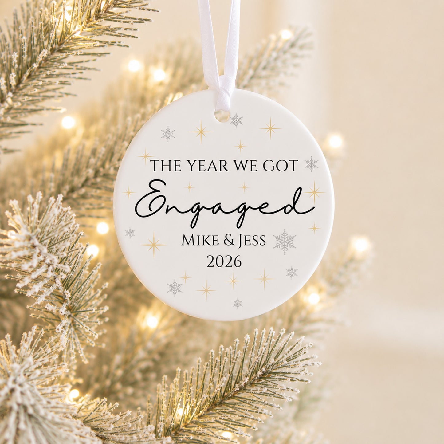 A round white Christmas ornament hanging from a white string, printed with the message 'THE YEAR WE GOT Engaged MIKE & JESS 2026' in black script and serif text, decorated with small gold and silver snowflake and star motifs. The ornament is photographed against a festive background of a lit flocked Christmas tree.