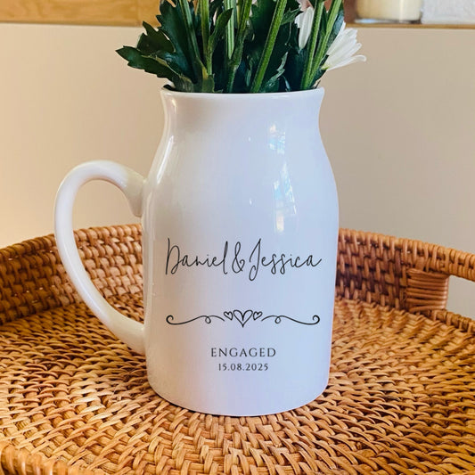 Personalised engagement vase. White pitcher with engraved text on a woven tray, surrounded by white flowers.