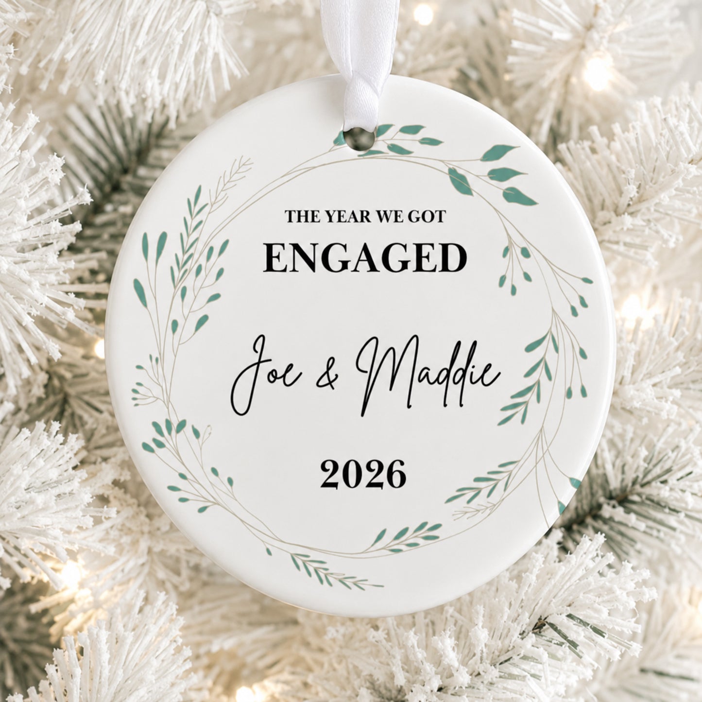A round white Christmas ornament with a hanging ribbon, printed with the text 'THE YEAR WE GOT ENGAGED Joe & Maddie 2026' inside a delicate green leaf wreath, displayed on a white flocked Christmas tree with warm string lights.