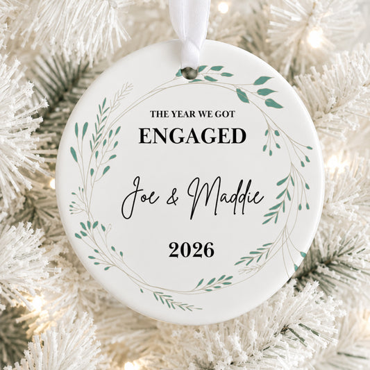 A round white Christmas ornament with a hanging ribbon, printed with the text 'THE YEAR WE GOT ENGAGED Joe & Maddie 2026' inside a delicate green leaf wreath, displayed on a white flocked Christmas tree with warm string lights.