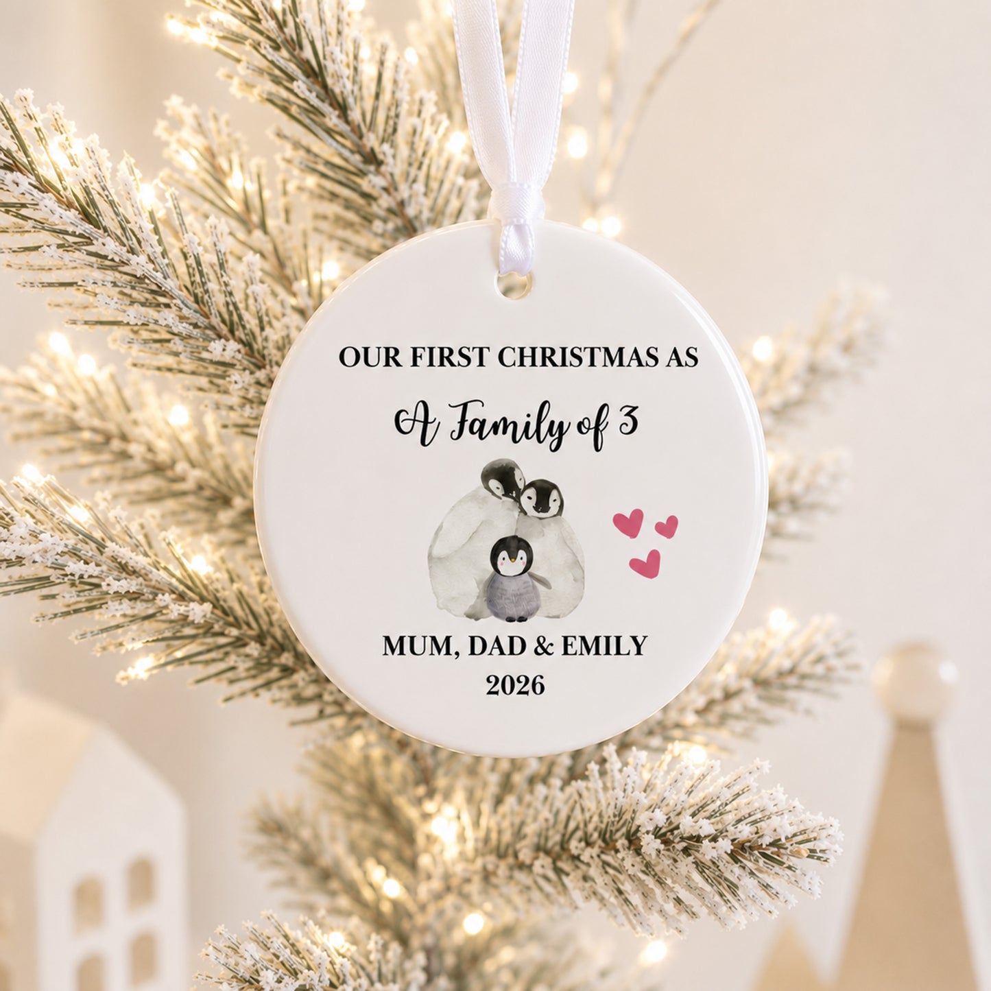 A personalized round white Christmas ornament hanging on a frosted tree with warm string lights. The ornament features black text reading 'OUR FIRST CHRISTMAS AS A Family of 3' and 'MUM, DAD & EMILY 2026', along with a small penguin illustration and pink hearts.