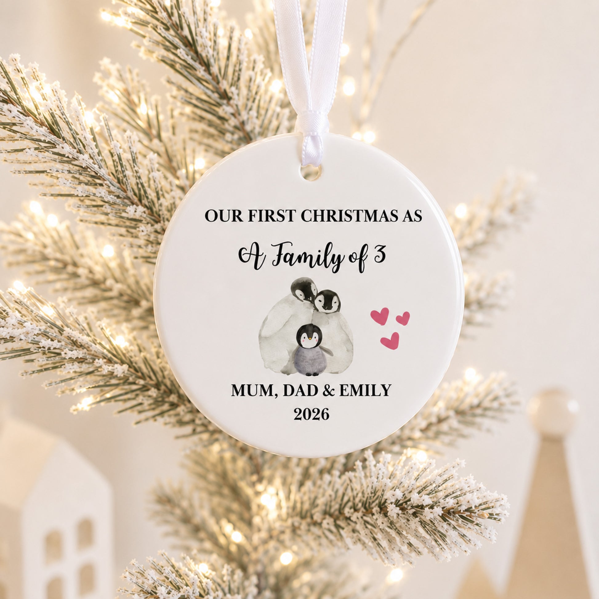 A personalized round white Christmas ornament hanging on a frosted tree with warm string lights. The ornament features black text reading 'OUR FIRST CHRISTMAS AS A Family of 3' and 'MUM, DAD & EMILY 2026', along with a small penguin illustration and pink hearts.