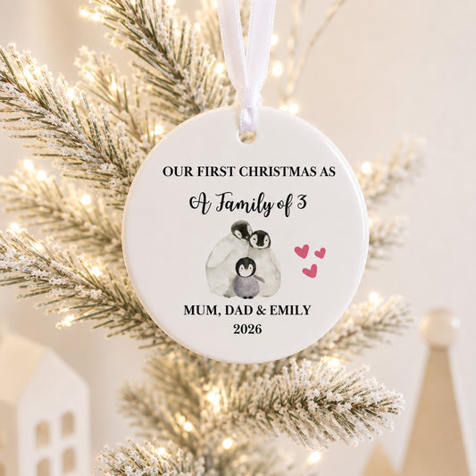 A personalized round white Christmas ornament hanging on a frosted tree with warm string lights. The ornament features black text reading 'OUR FIRST CHRISTMAS AS A Family of 3' and 'MUM, DAD & EMILY 2026', along with a small penguin illustration and pink hearts.