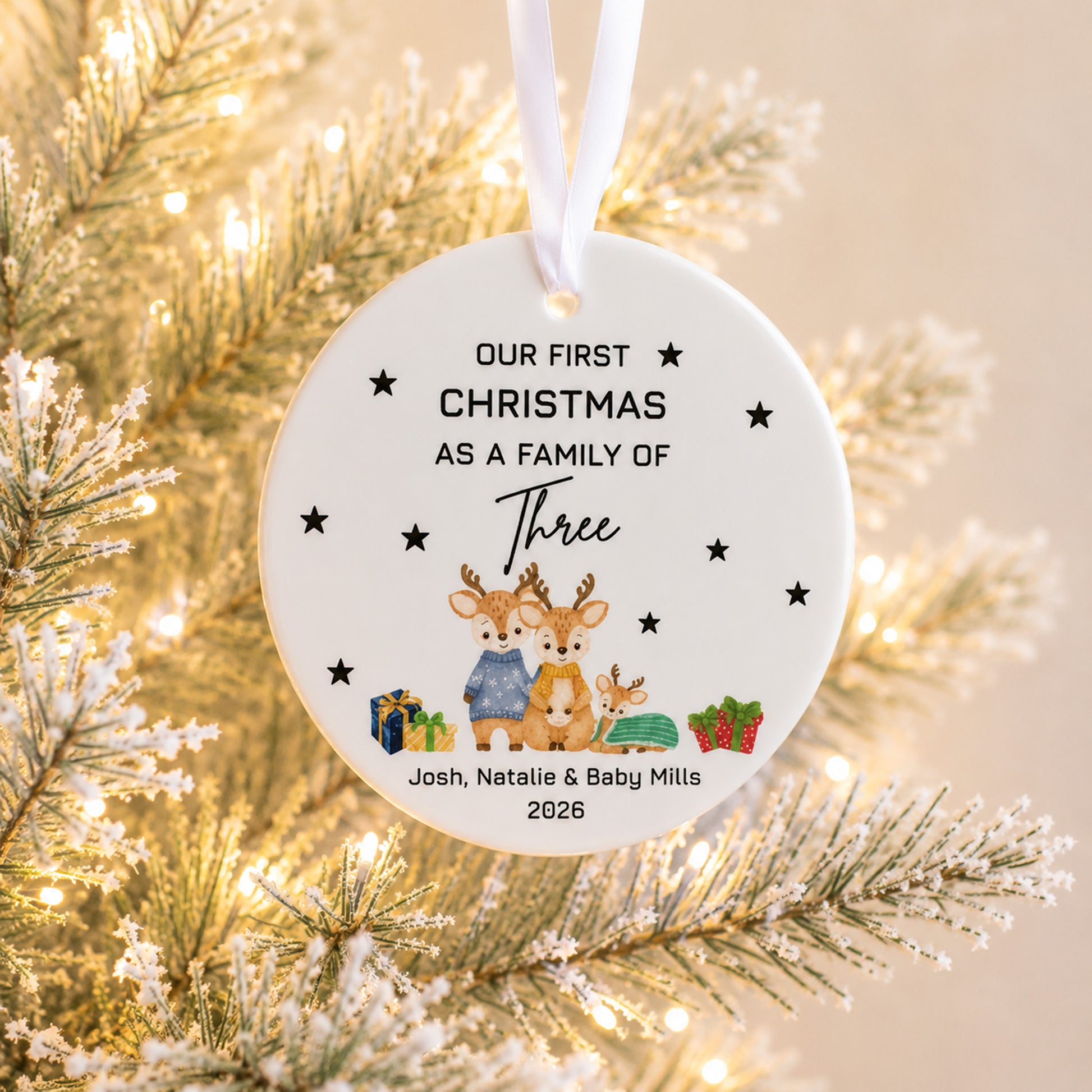 A personalized round Christmas ornament hanging from a white ribbon, featuring a watercolor illustration of three deer and gifts, with the text 'Our First Christmas As A Family Of Three Josh, Natalie & Baby Mills 2026'. The ornament is shown hanging in front of a lit, snowy pine tree.