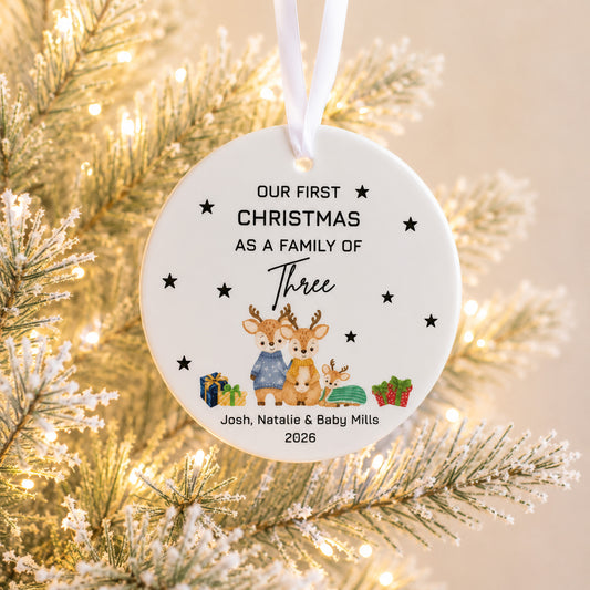 A personalized round Christmas ornament hanging from a white ribbon, featuring a watercolor illustration of three deer and gifts, with the text 'Our First Christmas As A Family Of Three Josh, Natalie & Baby Mills 2026'. The ornament is shown hanging in front of a lit, snowy pine tree.