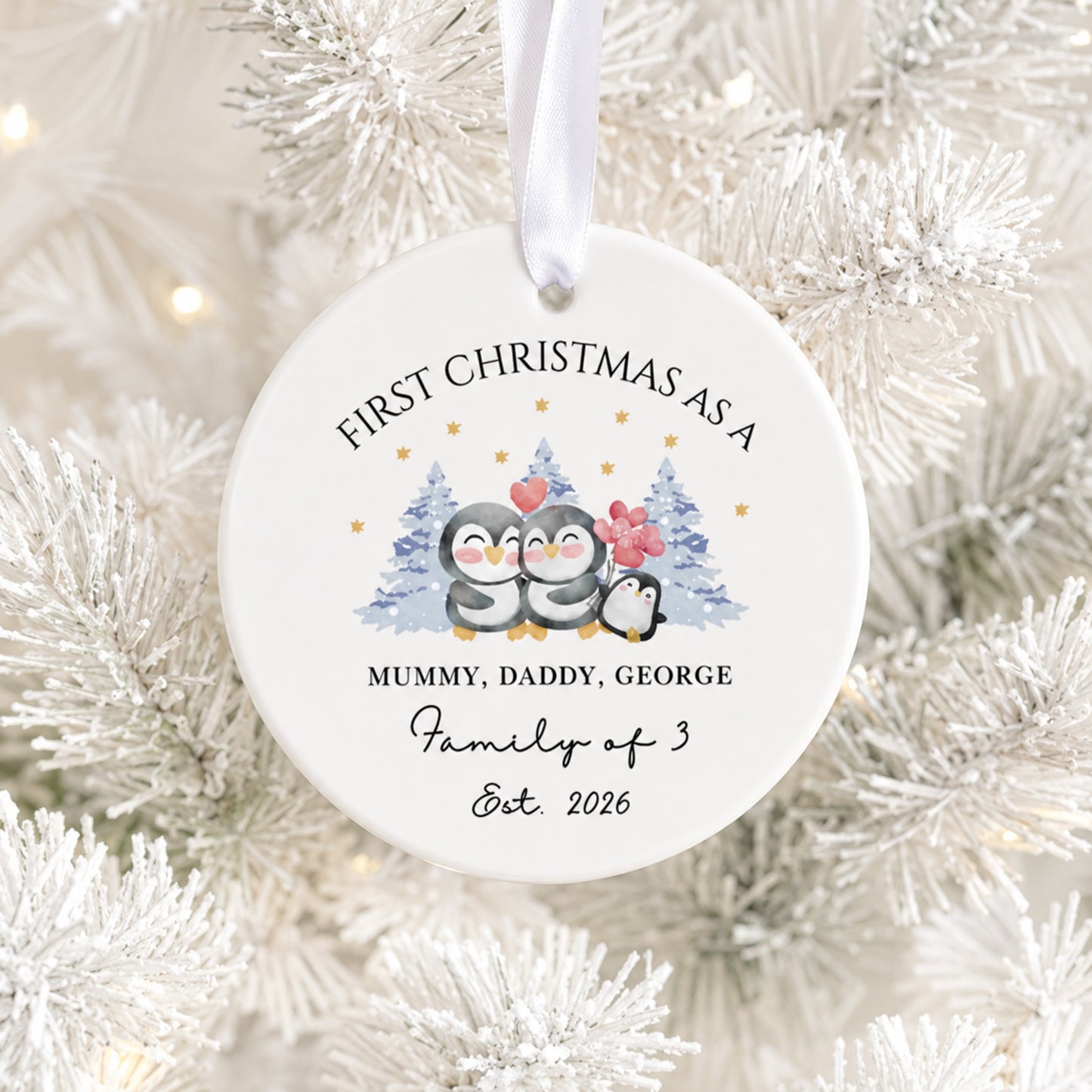 A round white ceramic Christmas ornament hanging on a white frosted Christmas tree. The ornament features a printed illustration of three penguins with small blue and red Christmas trees, along with personalized text reading 'FIRST CHRISTMAS AS A MUMMY, DADDY, GEORGE Family of 3 Est.