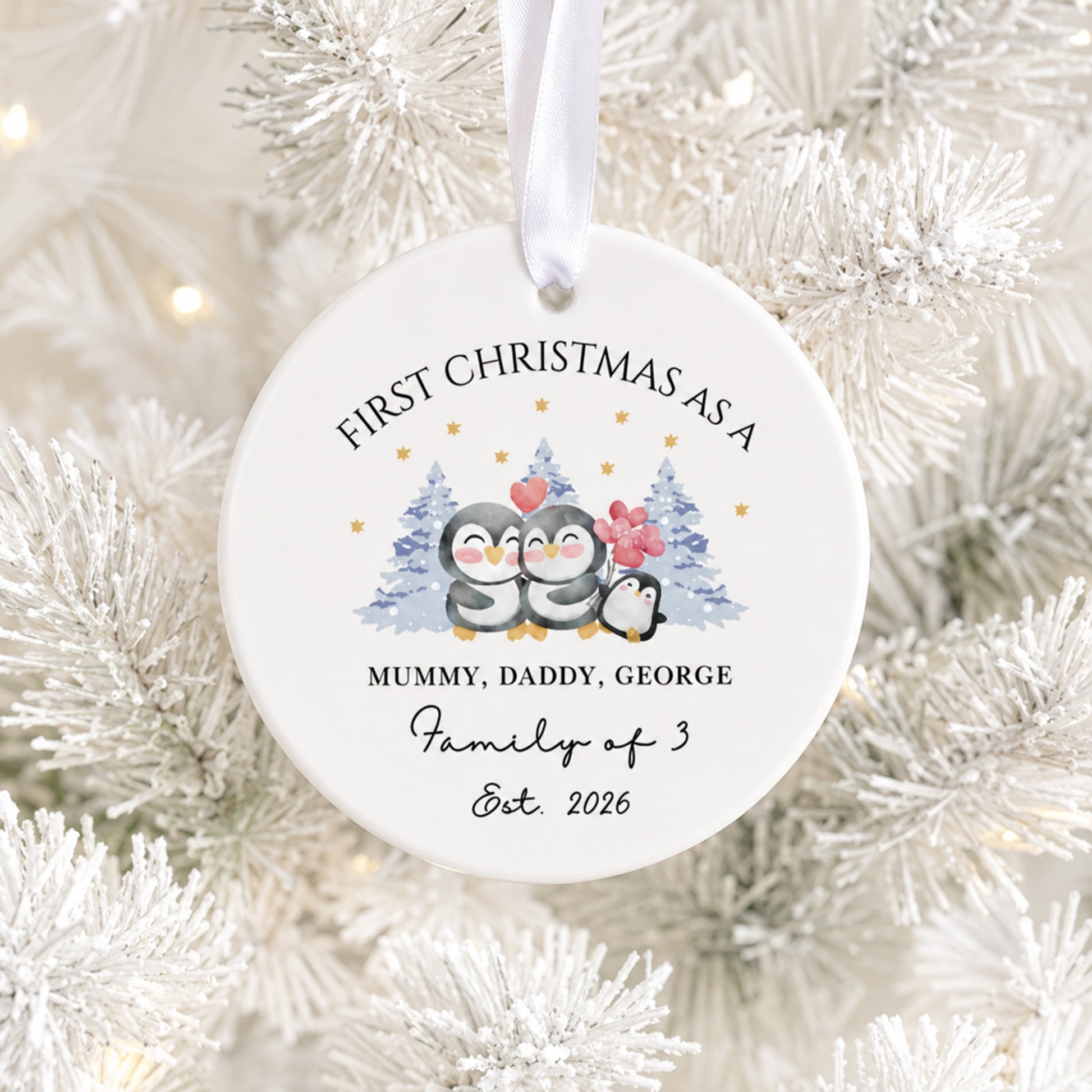 A round white ceramic Christmas ornament hanging on a white frosted Christmas tree. The ornament features a printed illustration of three penguins with small blue and red Christmas trees, along with personalized text reading 'FIRST CHRISTMAS AS A MUMMY, DADDY, GEORGE Family of 3 Est.