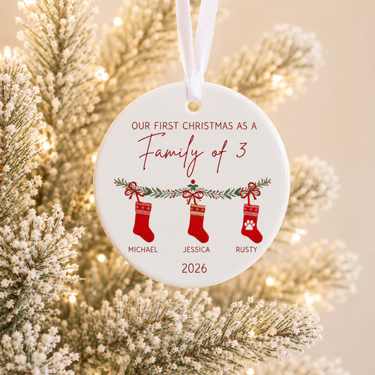 A round white personalized Christmas ornament hanging from a white ribbon, shown in front of a softly blurred frosted Christmas tree with warm lights. The ornament reads 'OUR FIRST CHRISTMAS AS A Family of 3' with three red stockings labeled Michael, Jessica, and Rusty, plus the year 2026.