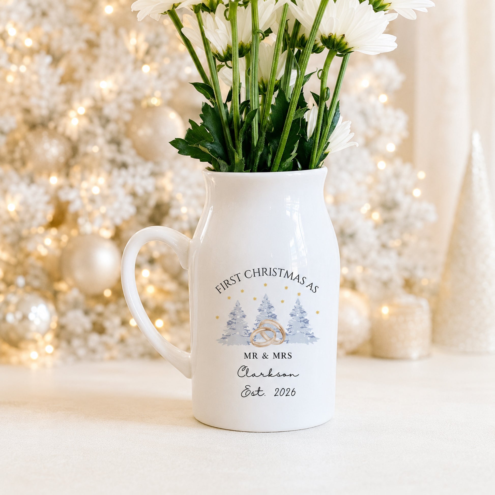 A white ceramic jug vase with a handle, filled with white daisy flowers, featuring a personalized Christmas mr and mrs design with a ring and trees. The background shows a softly blurred, festive white Christmas tree with warm lights.