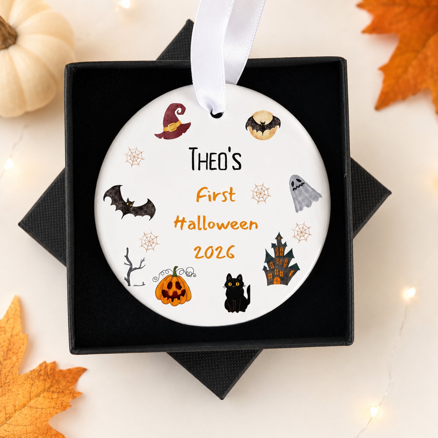 A personalized round white Christmas ornament with Halloween-themed illustrations and the text 'THEO'S First Halloween 2026', presented in a black gift box with a white ribbon, surrounded by autumn leaves and a small pumpkin.