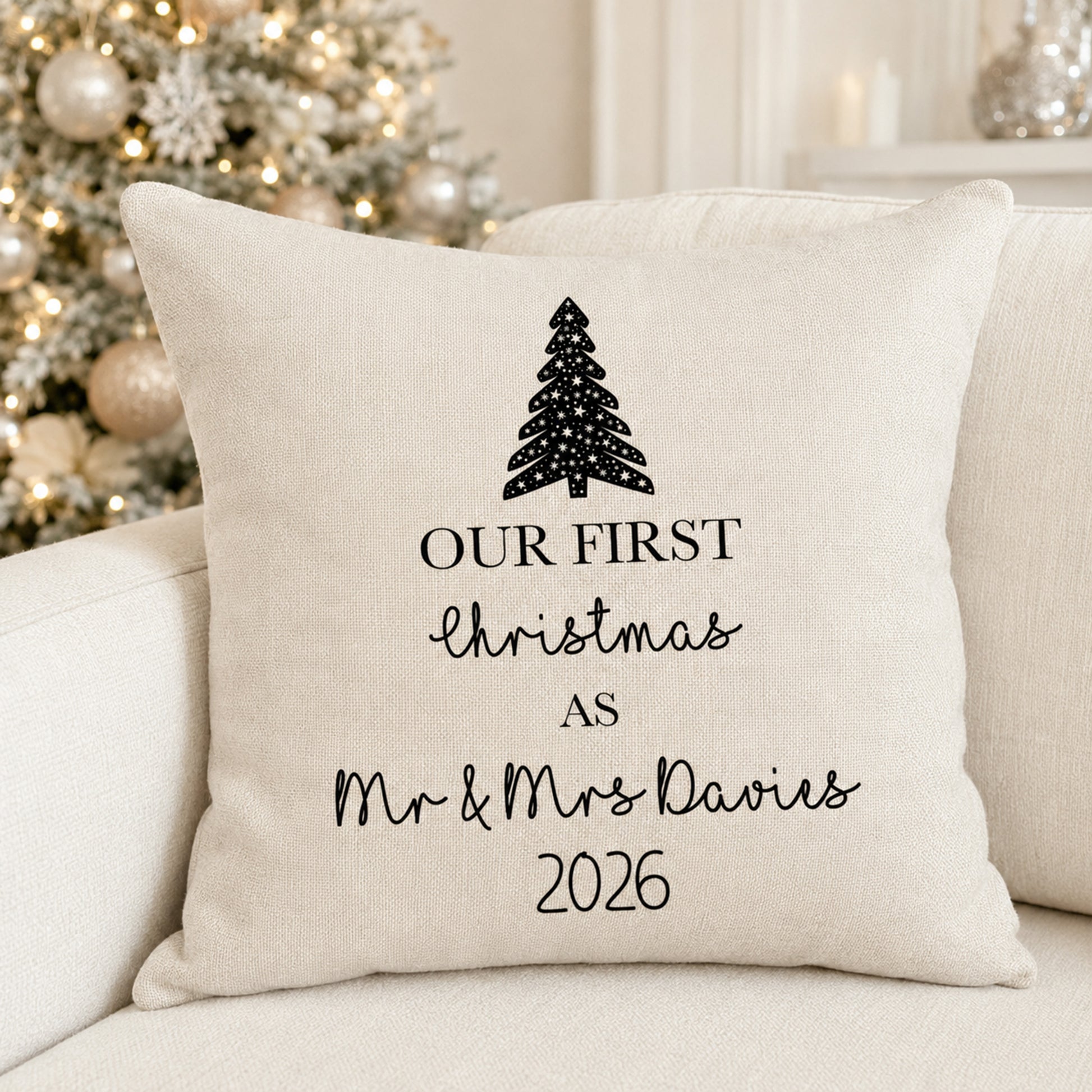 A square cream-colored throw pillow with black printed text reading 'OUR FIRST Christmas AS Mr & Mrs Davies 2026' and a black Christmas tree graphic, resting on a light beige sofa with a blurred decorated Christmas tree in the background.