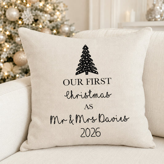 A square cream-colored throw pillow with black printed text reading 'OUR FIRST Christmas AS Mr & Mrs Davies 2026' and a black Christmas tree graphic, resting on a light beige sofa with a blurred decorated Christmas tree in the background.