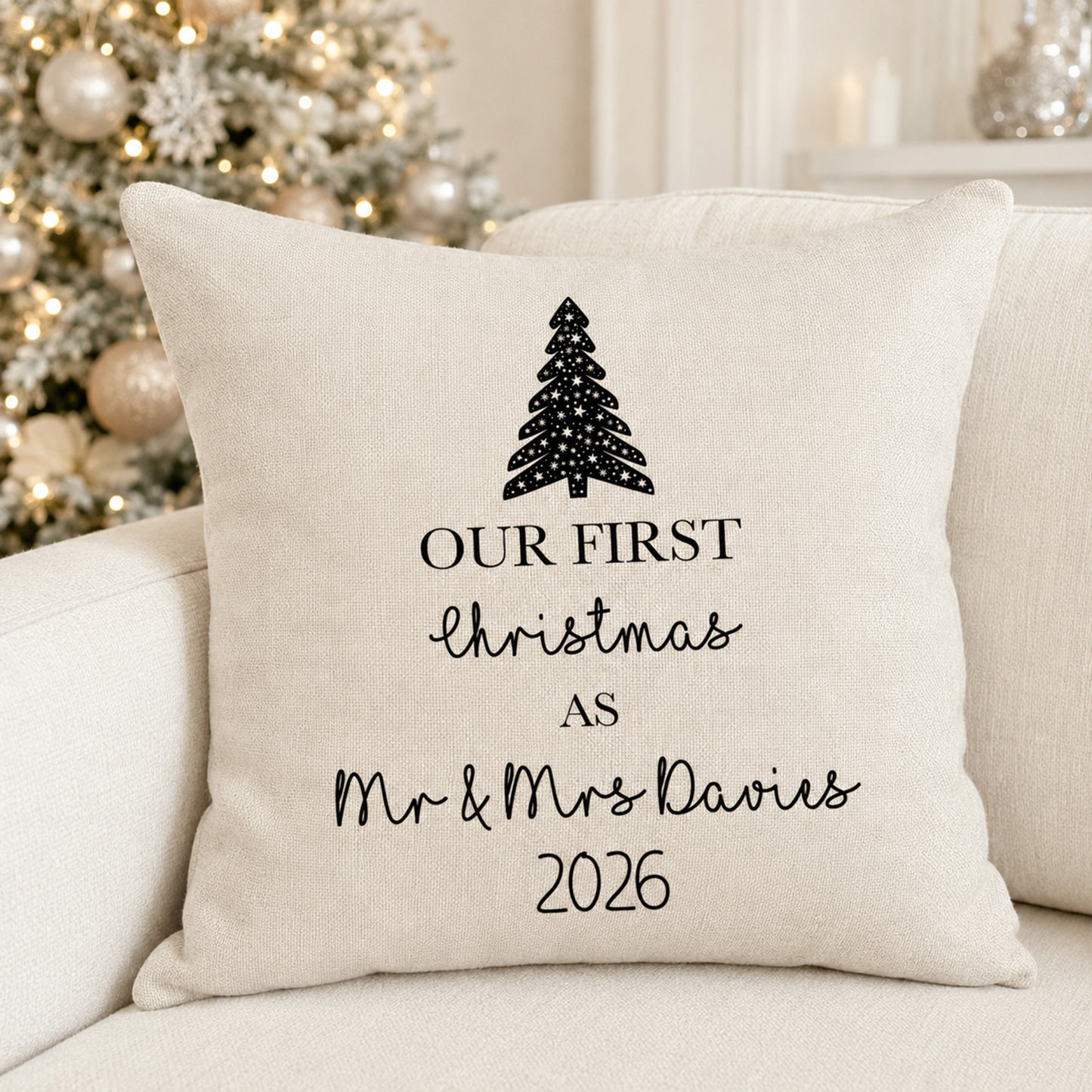 A square cream-colored throw pillow with black printed text reading 'OUR FIRST Christmas AS Mr & Mrs Davies 2026' and a black Christmas tree graphic, resting on a light beige sofa with a blurred decorated Christmas tree in the background.