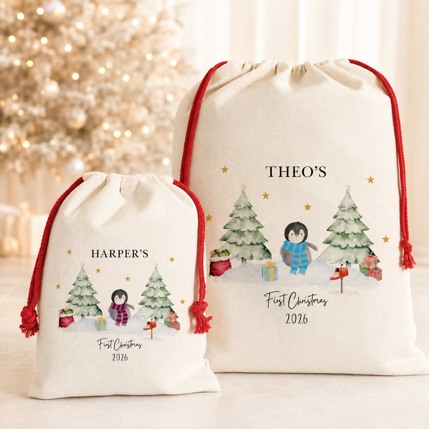 Two personalized first Christmas 2026 cream canvas drawstring gift bags with red cords, featuring winter penguin illustrations and text, set against a softly blurred holiday tree background.