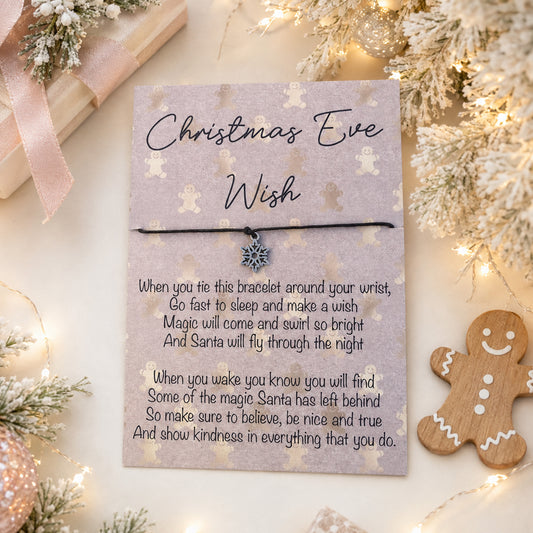 A Christmas-themed bracelet gift card featuring a black cord bracelet with a silver snowflake charm, displayed on a decorated Christmas Eve background with lights, pine branches, a wooden gingerbread cookie, and wrapped gifts.