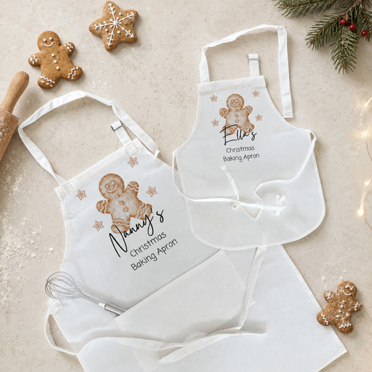 Two white Christmas baking aprons laid flat on a light beige surface, decorated with gingerbread man illustrations and the names 'Nancy's' and 'Ellis's Christmas Baking Apron.' The scene is styled with gingerbread cookies, a rolling pin, a whisk, and evergreen branches.