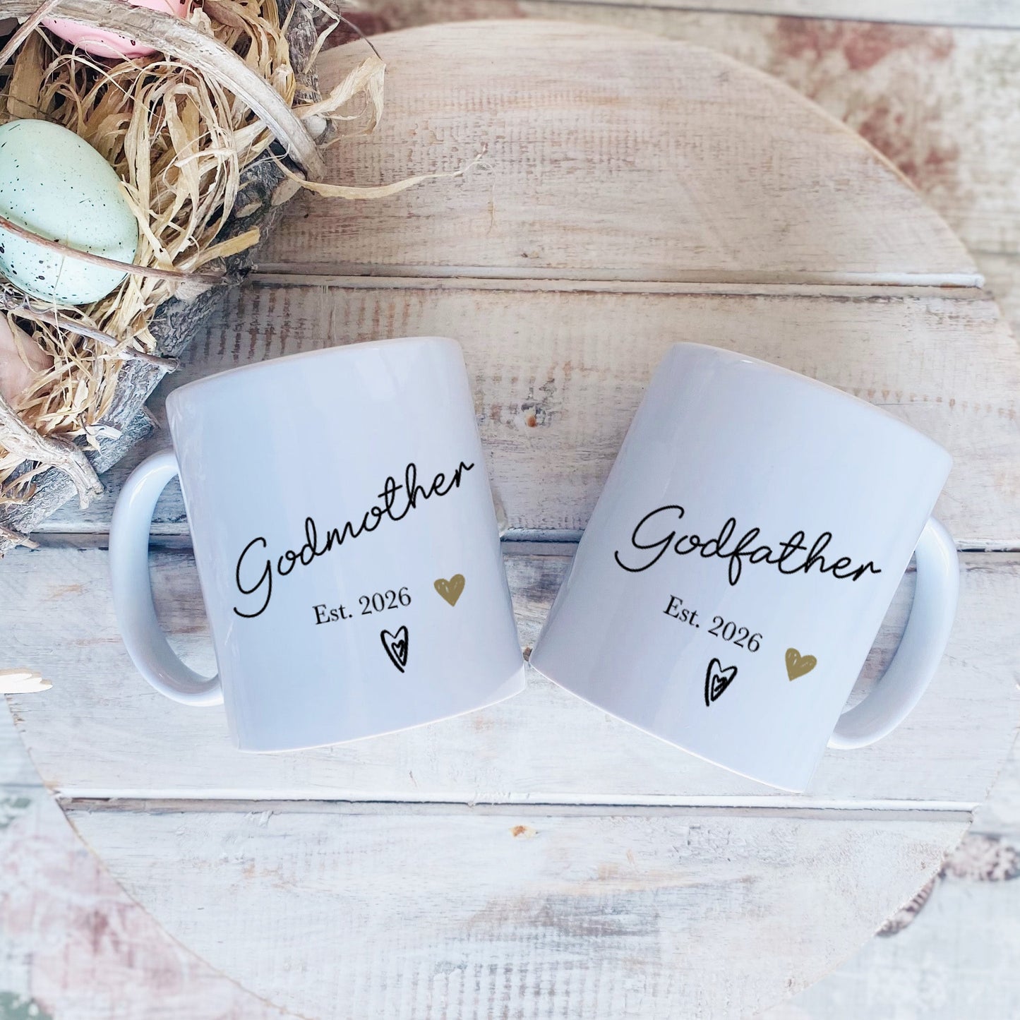 Two white mugs labeled 'Godmother' and 'Godfather' with a decorative nest and eggs on a wooden surface.