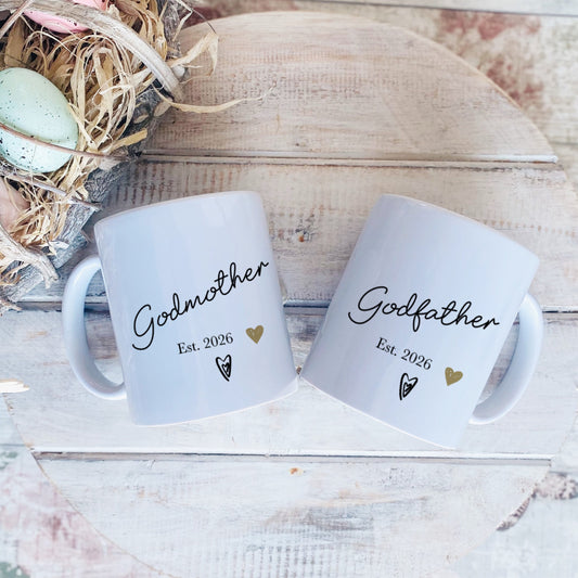 Two white mugs labeled 'Godmother' and 'Godfather' with a decorative nest and eggs on a wooden surface.