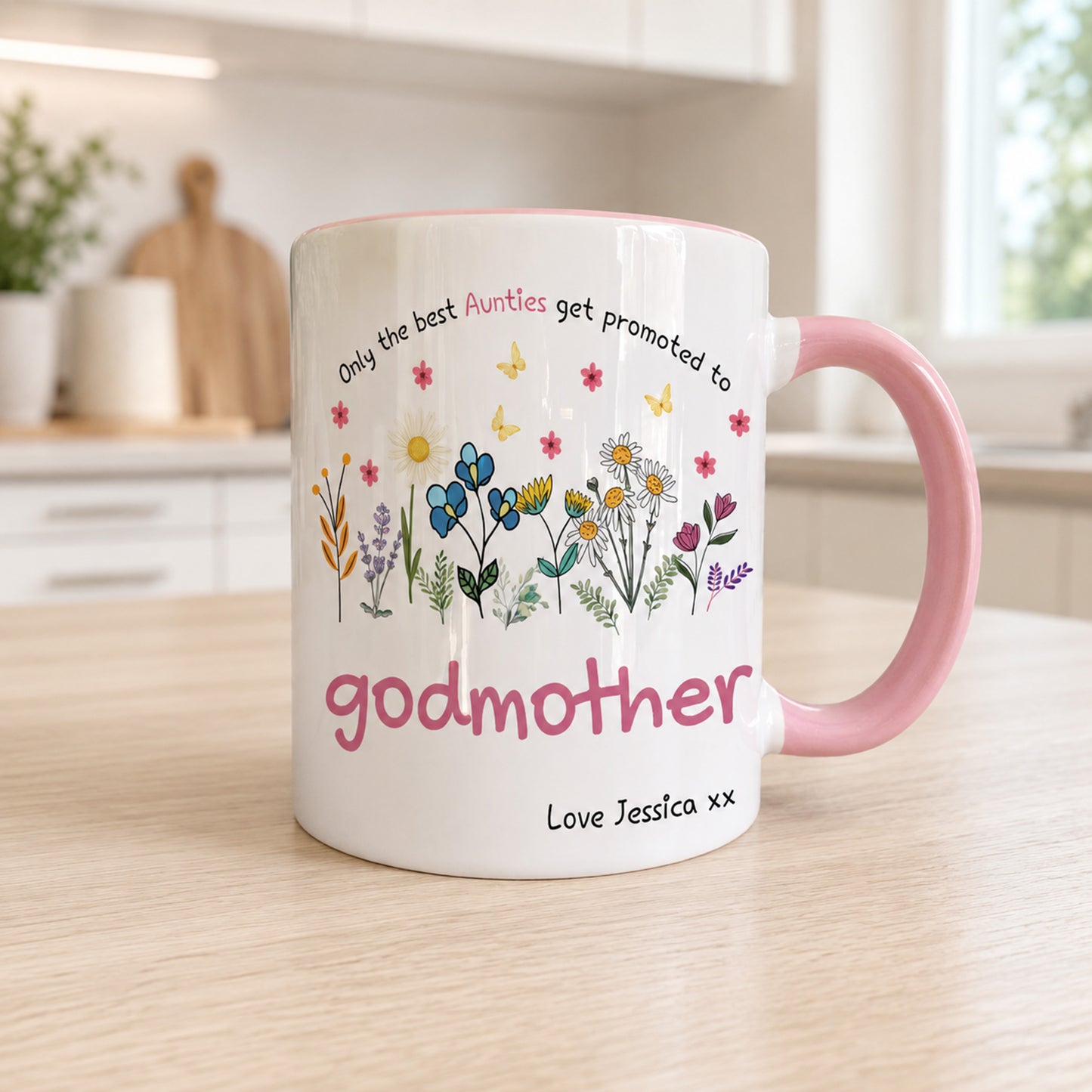 A white ceramic mug with a pink handle and pink interior, decorated with colorful wildflowers, butterflies, and the text 'Only the best Aunties get promoted to godmother' and 'Love Jessica xx'. The mug is shown on a light wood tabletop in a softly blurred kitchen setting.