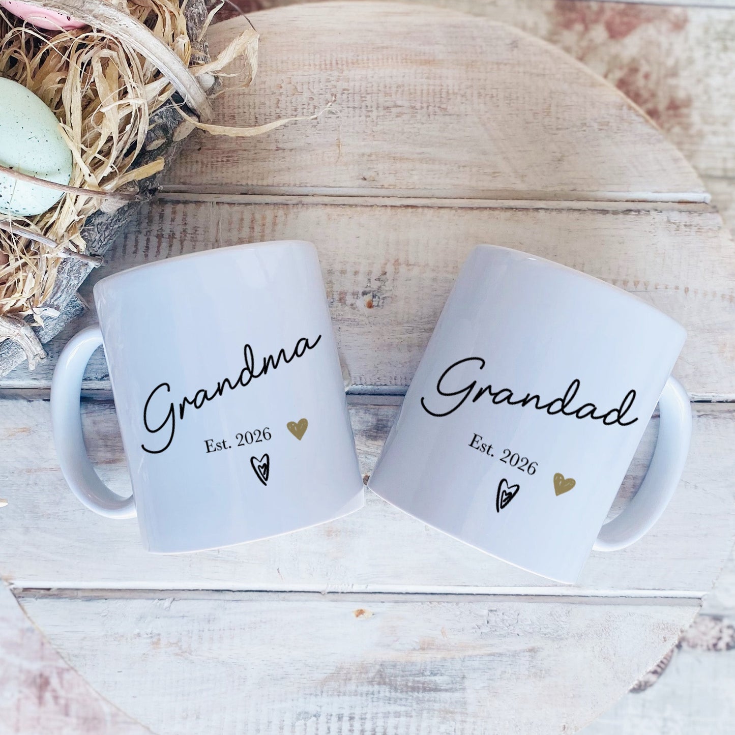 Two white mugs with 'Grandma' and 'Grandad' text on a rustic wooden surface with Easter eggs and nest.
