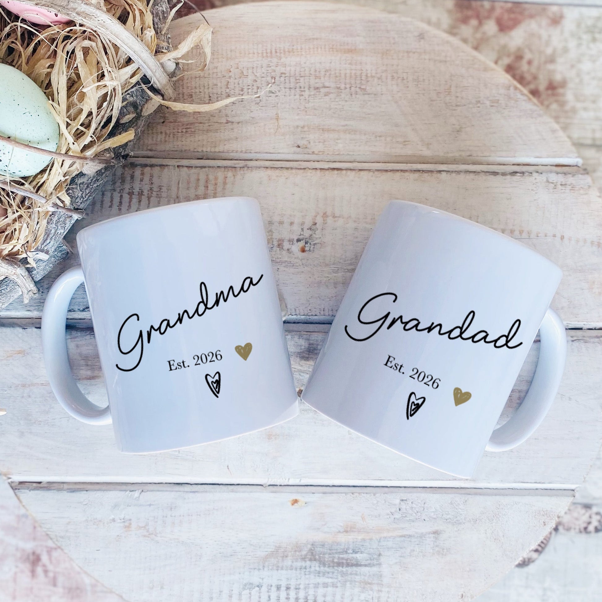 Two white mugs with 'Grandma' and 'Grandad' text on a rustic wooden surface with Easter eggs and nest.