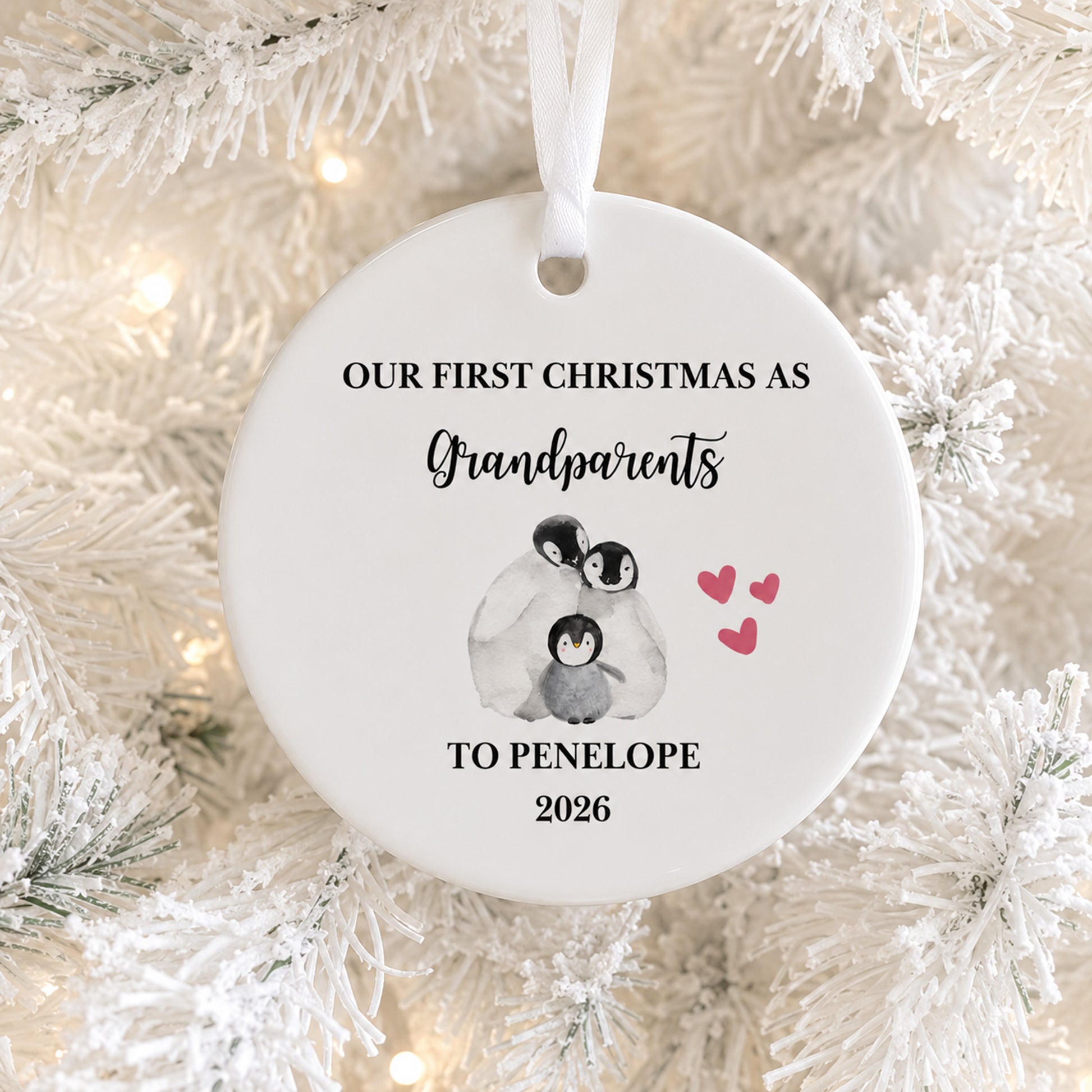 A round white ceramic Christmas ornament hanging on a white frosted tree, printed with the text 'OUR FIRST CHRISTMAS AS Grandparents TO PENELOPE 2026' and a small penguin illustration with pink hearts.