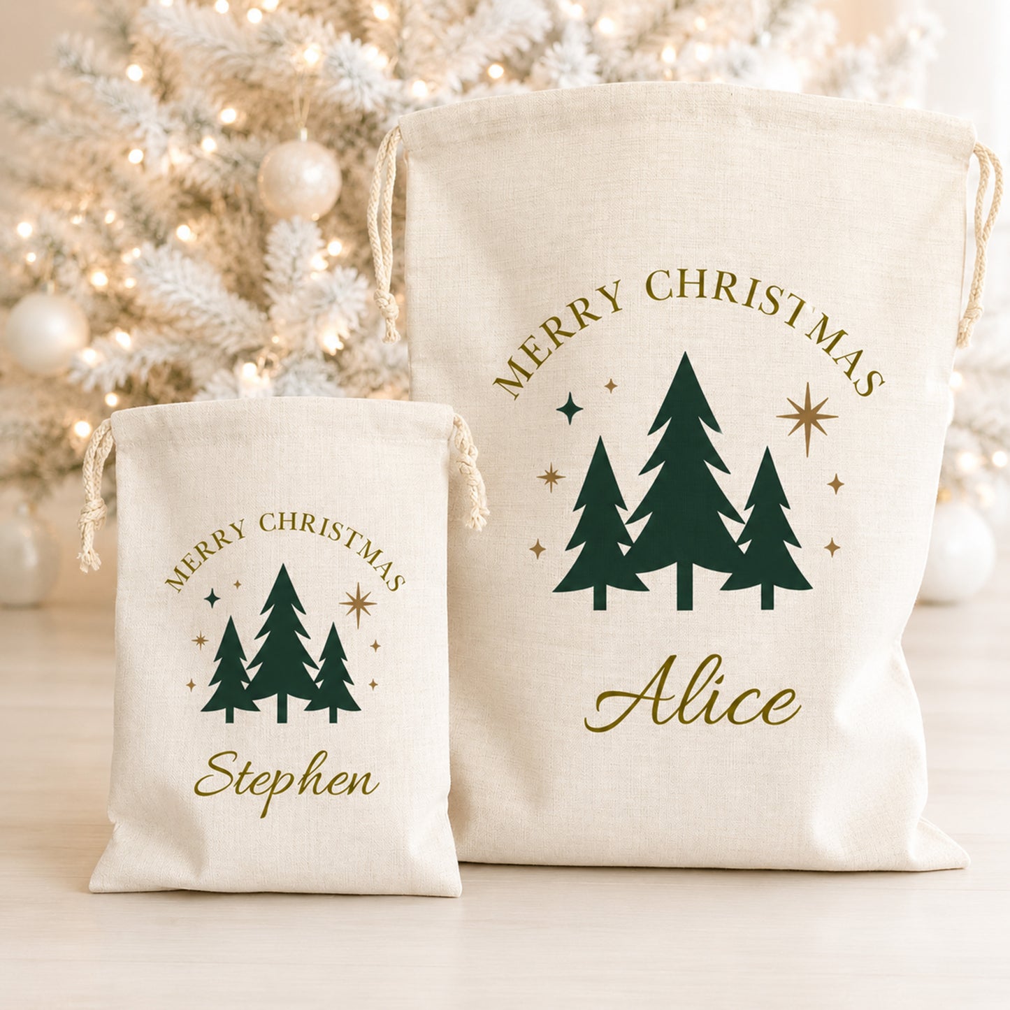 Two personalized canvas Christmas gift bags with drawstrings, one small and one large, featuring dark green pine tree graphics and gold text reading 'Merry Christmas' and the names 'Alice' and 'Stephen'. The bags are displayed in front of a softly blurred white Christmas tree with warm lights.