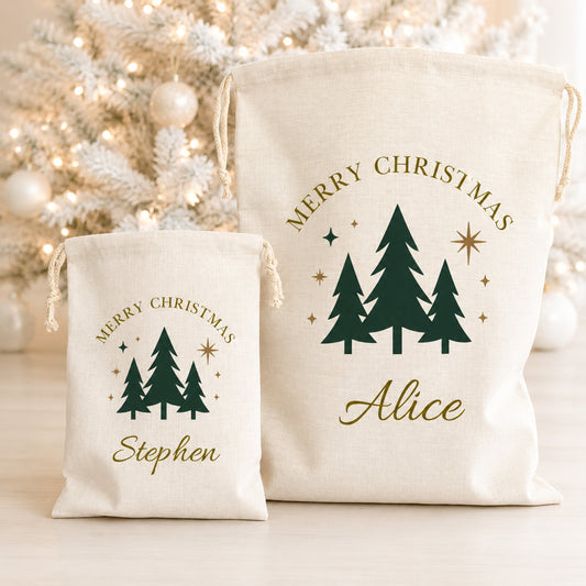 Two personalized canvas Christmas gift bags with drawstrings, one small and one large, featuring dark green pine tree graphics and gold text reading 'Merry Christmas' and the names 'Alice' and 'Stephen'. The bags are displayed in front of a softly blurred white Christmas tree with warm lights.