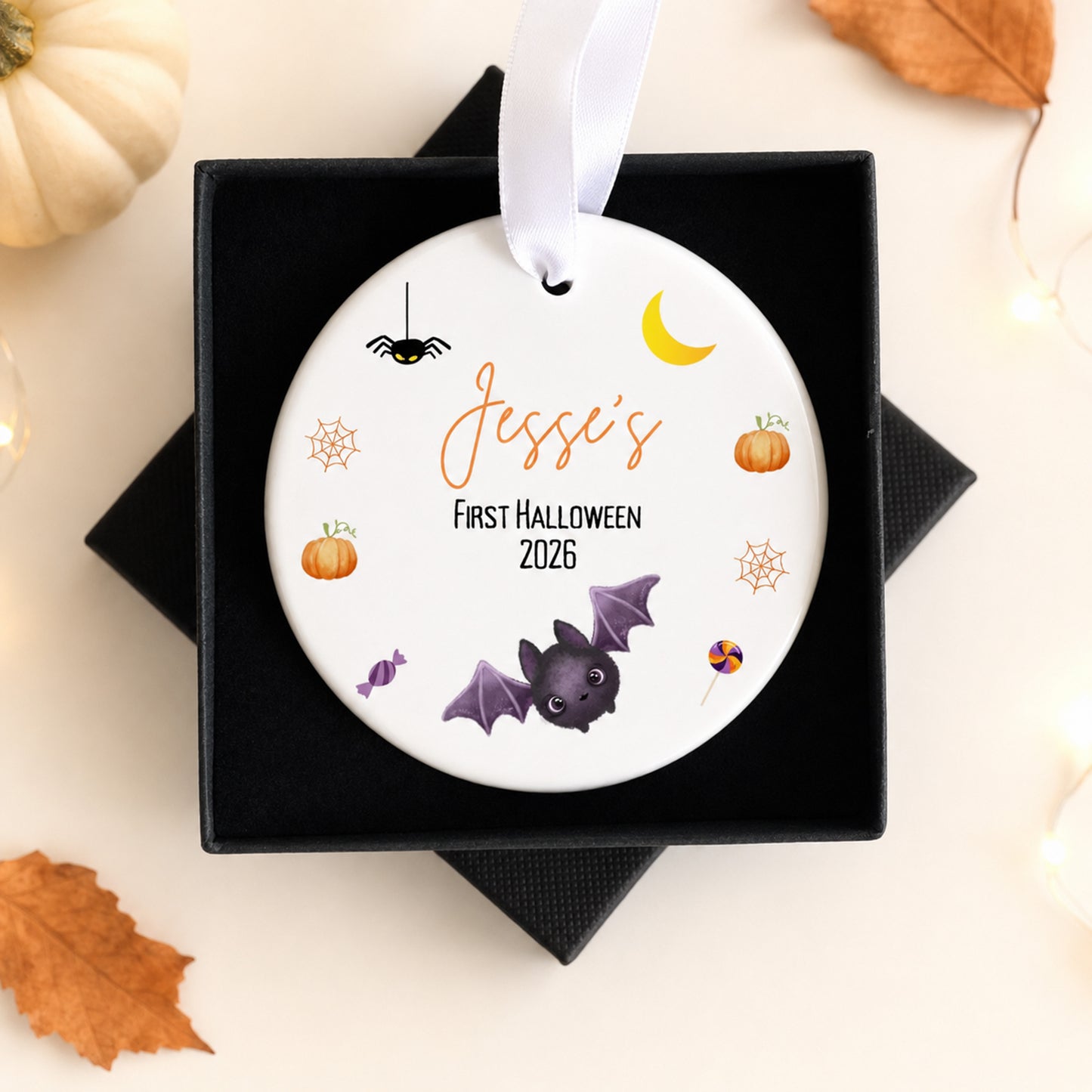 A round white personalized Halloween ornament with a white ribbon, displayed in a black gift box. The ornament features the text 'Jessie's First Halloween 2026' in orange and black, surrounded by cute illustrations of a purple bat, spider, pumpkins, spiderwebs, and a yellow crescent moon.