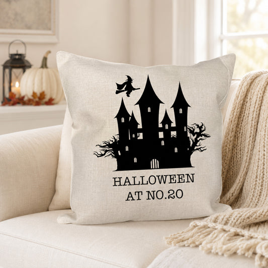 A square off-white throw pillow with a black haunted house graphic and the text 'HALLOWEEN AT NO.20' sits on a cream couch in a cozy living room setting with a knit blanket and pumpkins in the background.