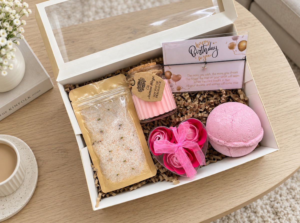 A curated gift box set on a light wood table, featuring a pink bath bomb, a candle, a clear bag of bath salts, and a pink birthday wish bracelet card, all arranged in a box with shredded paper filler.