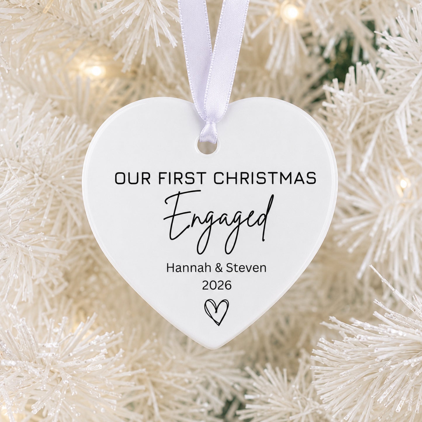 A white heart-shaped Christmas ornament hanging from a white ribbon, printed with the text 'OUR FIRST CHRISTMAS Engaged Hannah & Steven 2026' and a small heart, shown against a blurred white Christmas tree background with warm lights.