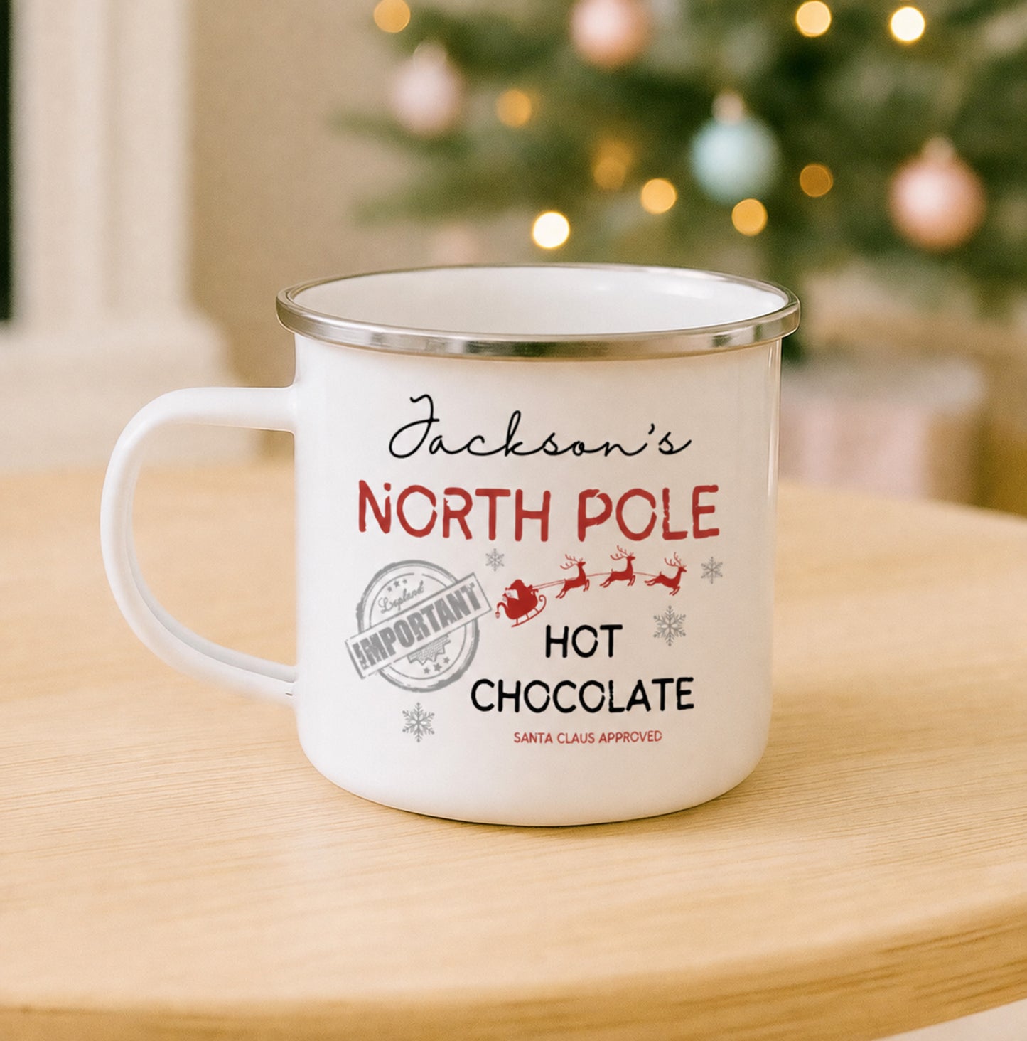 A white enamel-style mug with a silver rim sits on a light wood table in front of a blurred Christmas tree with warm lights. The mug is printed with the phrase 'Jackson's NORTH POLE HOT CHOCOLATE' in black and red lettering, along with small Santa and snowflake graphics.