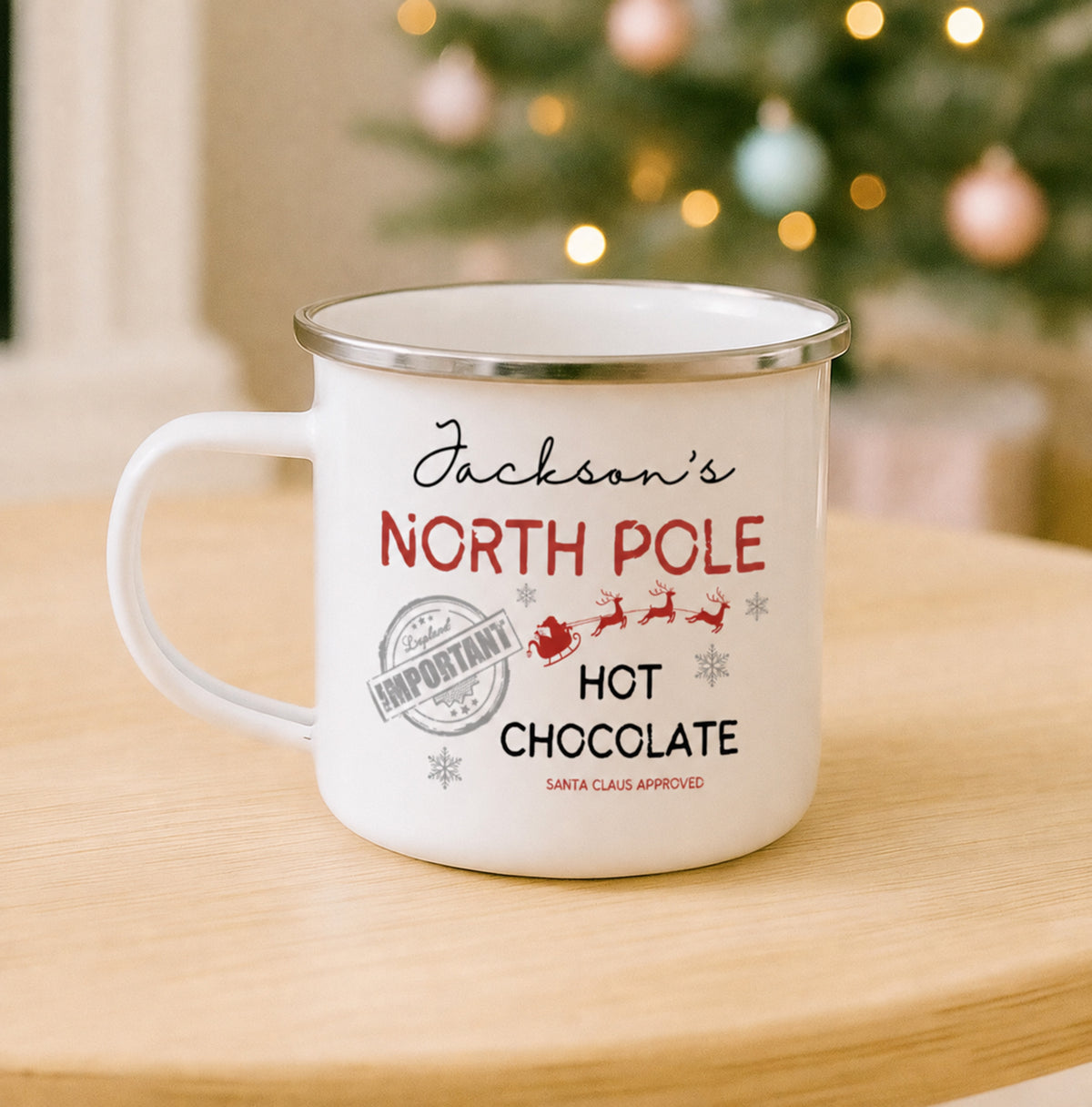 A white enamel-style mug with a silver rim sits on a light wood table in front of a blurred Christmas tree with warm lights. The mug is printed with the phrase 'Jackson's NORTH POLE HOT CHOCOLATE' in black and red lettering, along with small Santa and snowflake graphics.