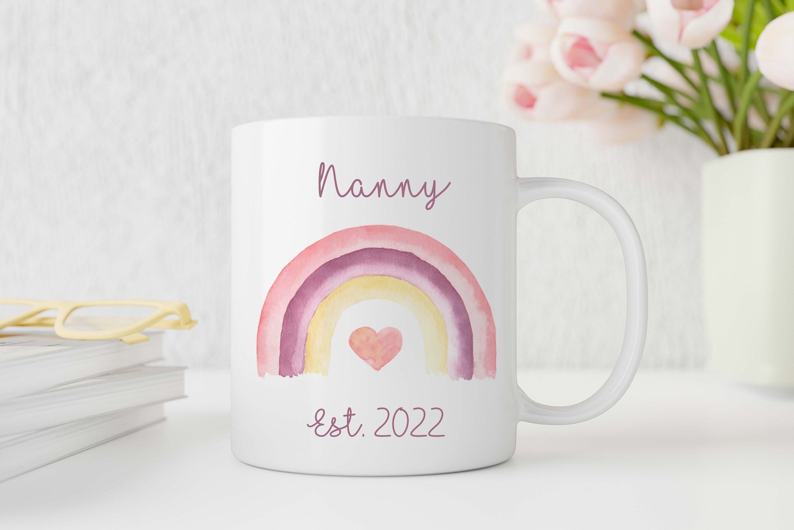 White mug with a rainbow design and 'Nanny Est. 2022' text on a white surface with flowers in the background.