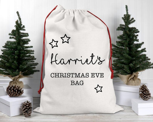 Personalised White drawstring bag labeled 'Harriet's Christmas Eve Bag' with decorative stars, placed between two small Christmas trees.