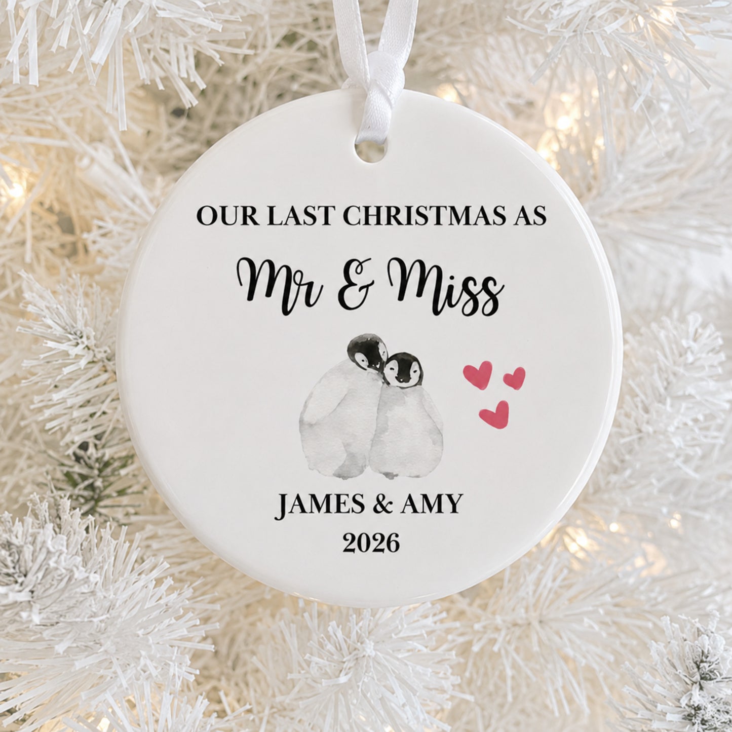 A round white ceramic Christmas ornament hanging on a white flocked Christmas tree with warm string lights. The ornament is personalized with the text 'OUR LAST CHRISTMAS AS Mr & Miss JAMES & AMY 2026' and features a watercolor illustration of two penguins with three small red hearts.