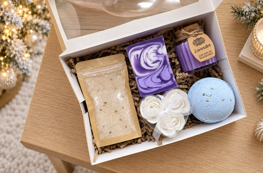 A curated bath and body gift box set displayed in a white box with a clear lid, filled with brown paper shred. The set includes a lavender soap bar, a blue bath bomb, a white rose-shaped item, a clear pouch of salt, and a small candle jar labeled Lavender.