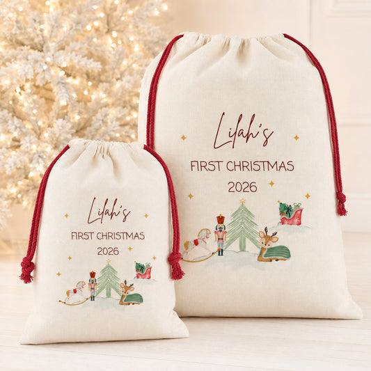 Two personalized cream drawstring gift bags with red cords, featuring a Christmas tree, nutcracker, reindeer, and sleigh illustration with the text 'Lilah's FIRST CHRISTMAS 2026', shown in front of a softly blurred white Christmas tree.